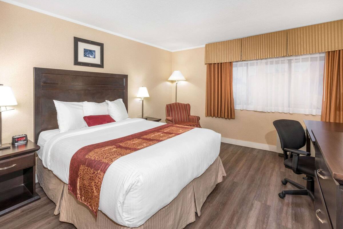 Ein Hotelzimmer mit einem großen Bett, zwei Nachttischlampen, einem Schreibtisch mit Stuhl, einem Fenster mit Vorhängen und einem gerahmten Bild an der Wand.