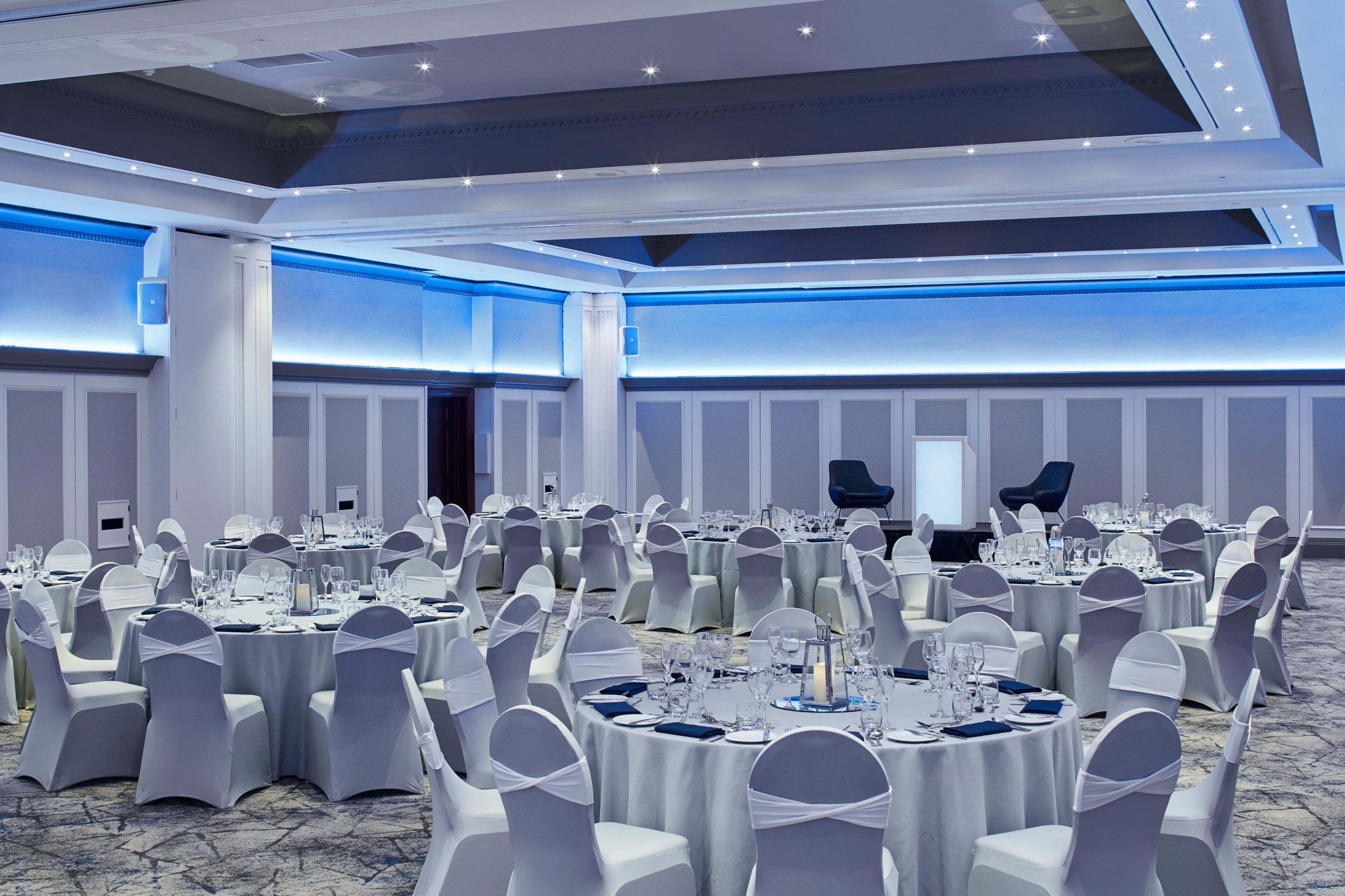A large banquet hall with round tables set for dining, white chairs, and a modern ceiling design.