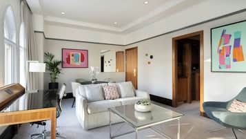 Ein modernes Wohnzimmer mit Glastisch, Sofa, Schreibtisch und einem Sessel. Ein großes Fenster mit Vorhängen, eine Pflanze und Kunstwerke an den Wänden.