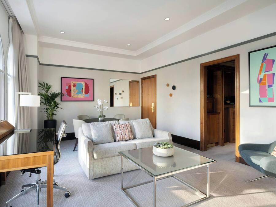 A modern living room with a glass coffee table, a sofa, a desk, and a chair. There is a large window with curtains, a plant, and artwork on the walls.