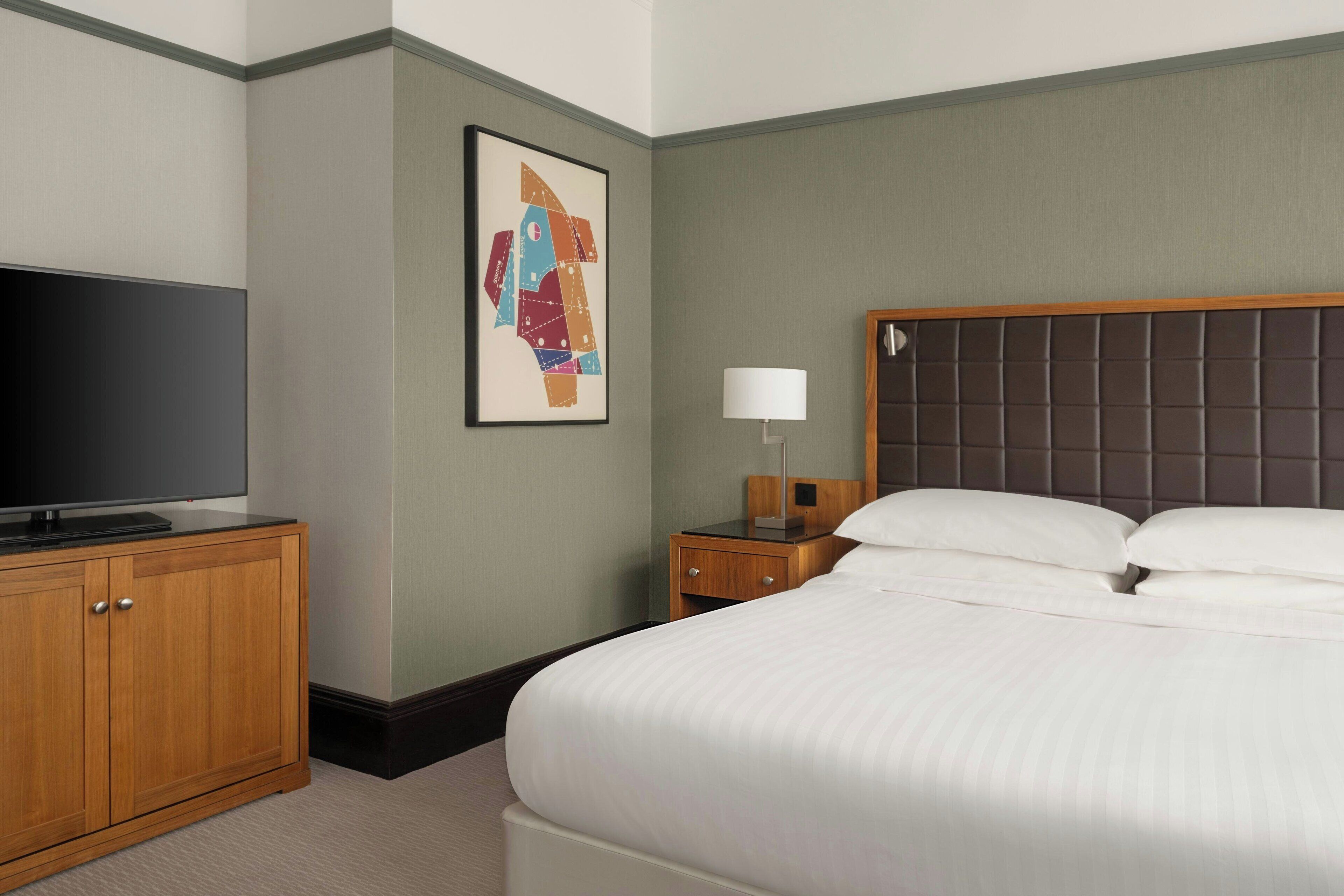 A hotel room with a bed, a television on a wooden cabinet, a nightstand with a lamp, and a framed abstract artwork on the wall.