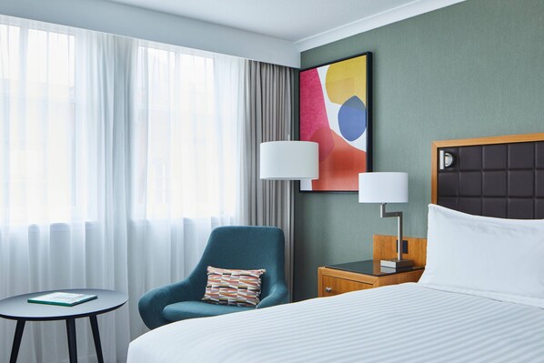 A hotel room with a bed, a chair, a small table, a lamp, and a colorful abstract painting on the wall.