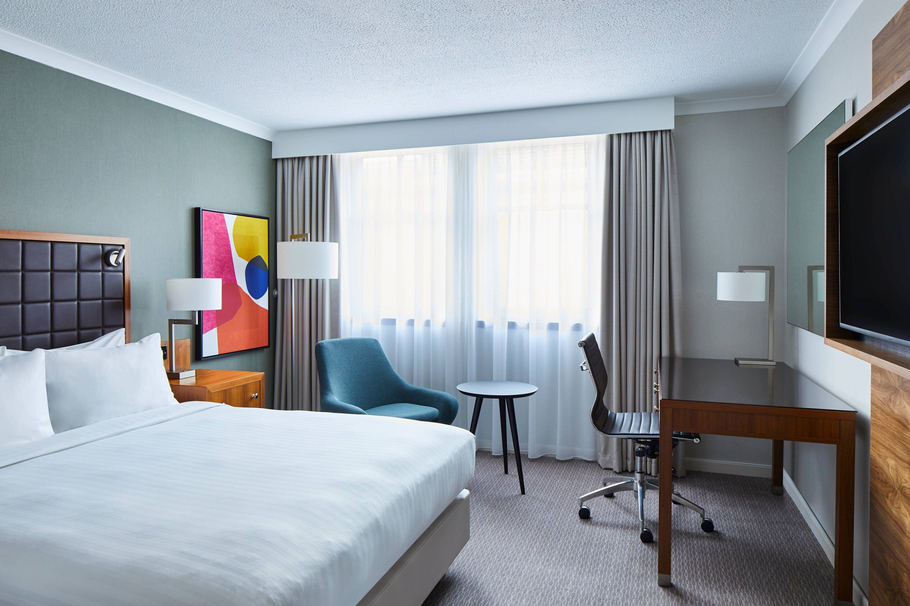 A hotel room with a large bed, a desk, a chair, a TV, and a window with curtains.