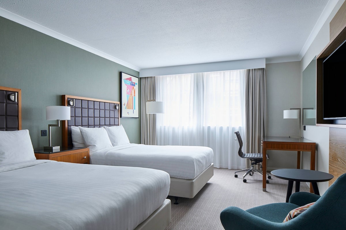 A hotel room with two beds, a desk, a chair, a TV, and a window with curtains.