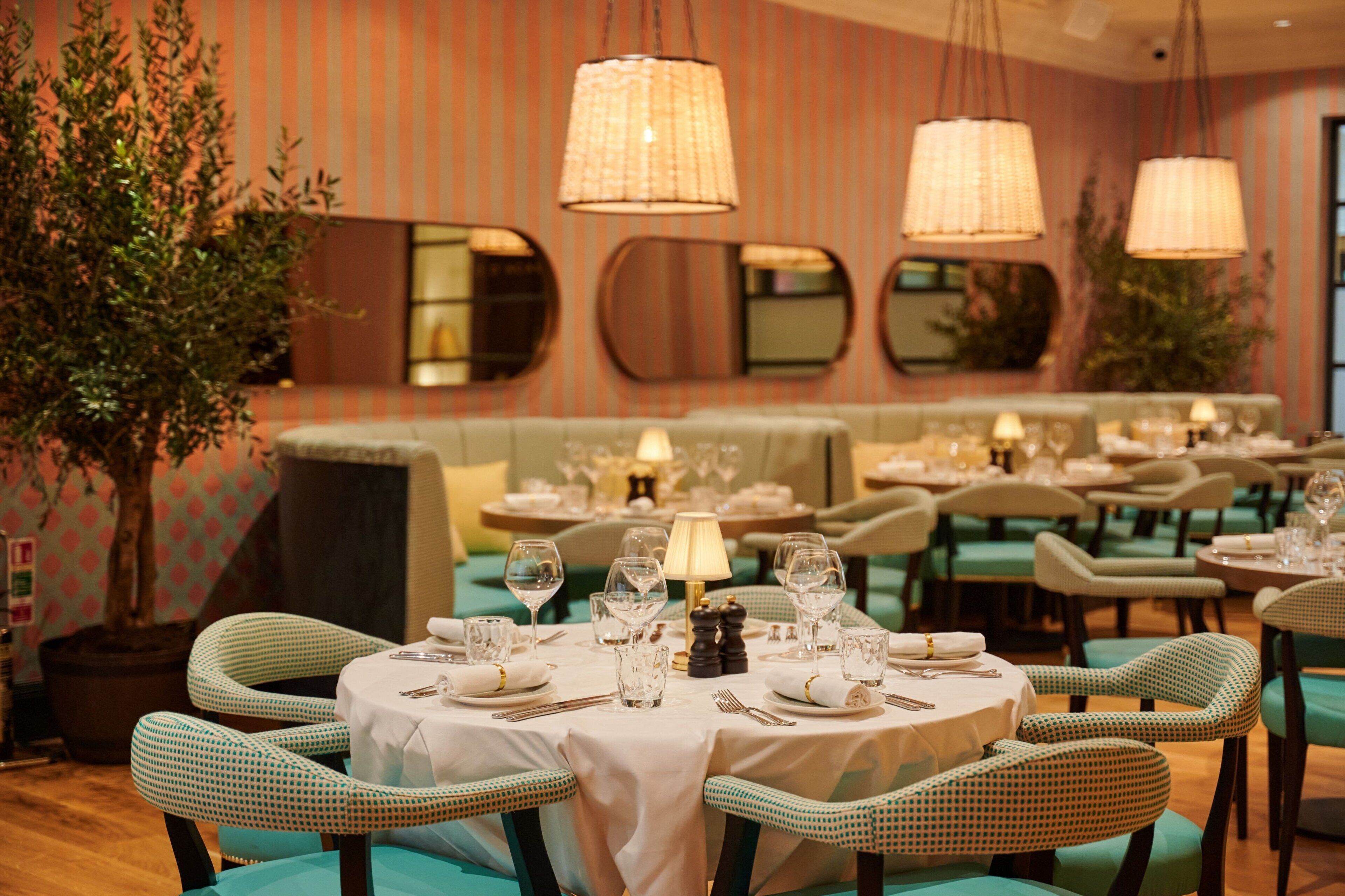 A restaurant with round tables set for dining, green upholstered chairs, and hanging lamps.