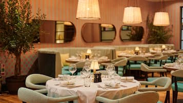 A restaurant with round tables set for dining, green upholstered chairs, and hanging lamps.