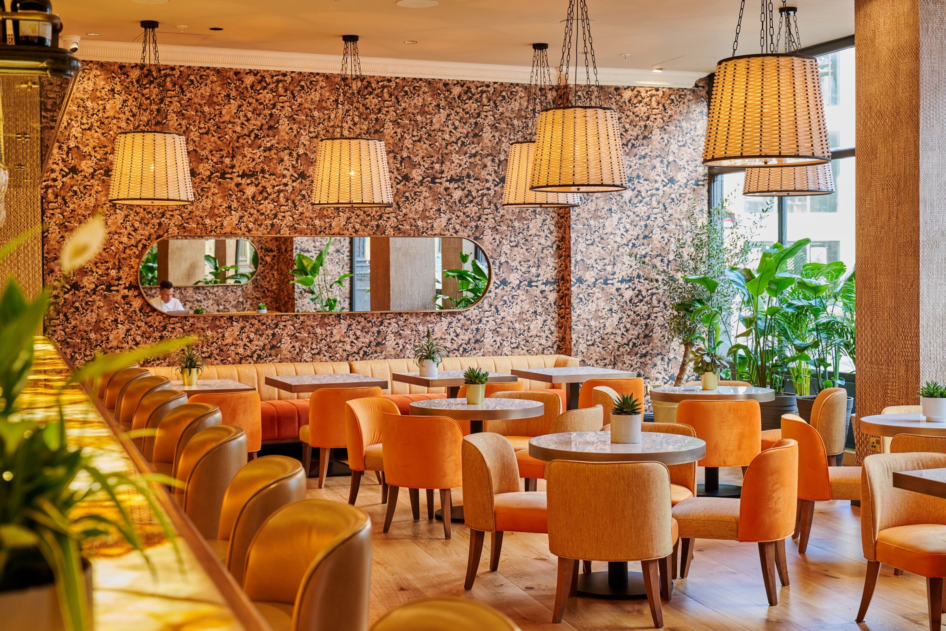 Un restaurante con mesas redondas y sillas naranjas, una pared de piedra con espejos, y lámparas de mimbre colgantes.
