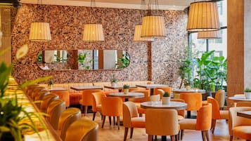 Un restaurante con mesas redondas y sillas naranjas, una pared de piedra con espejos, y lámparas de mimbre colgantes.