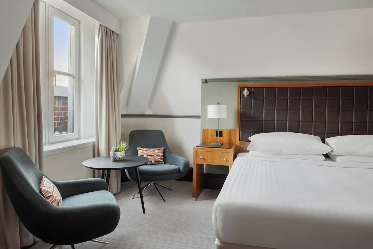 A hotel room with a bed, a chair, a small table, a nightstand, and a window with curtains.