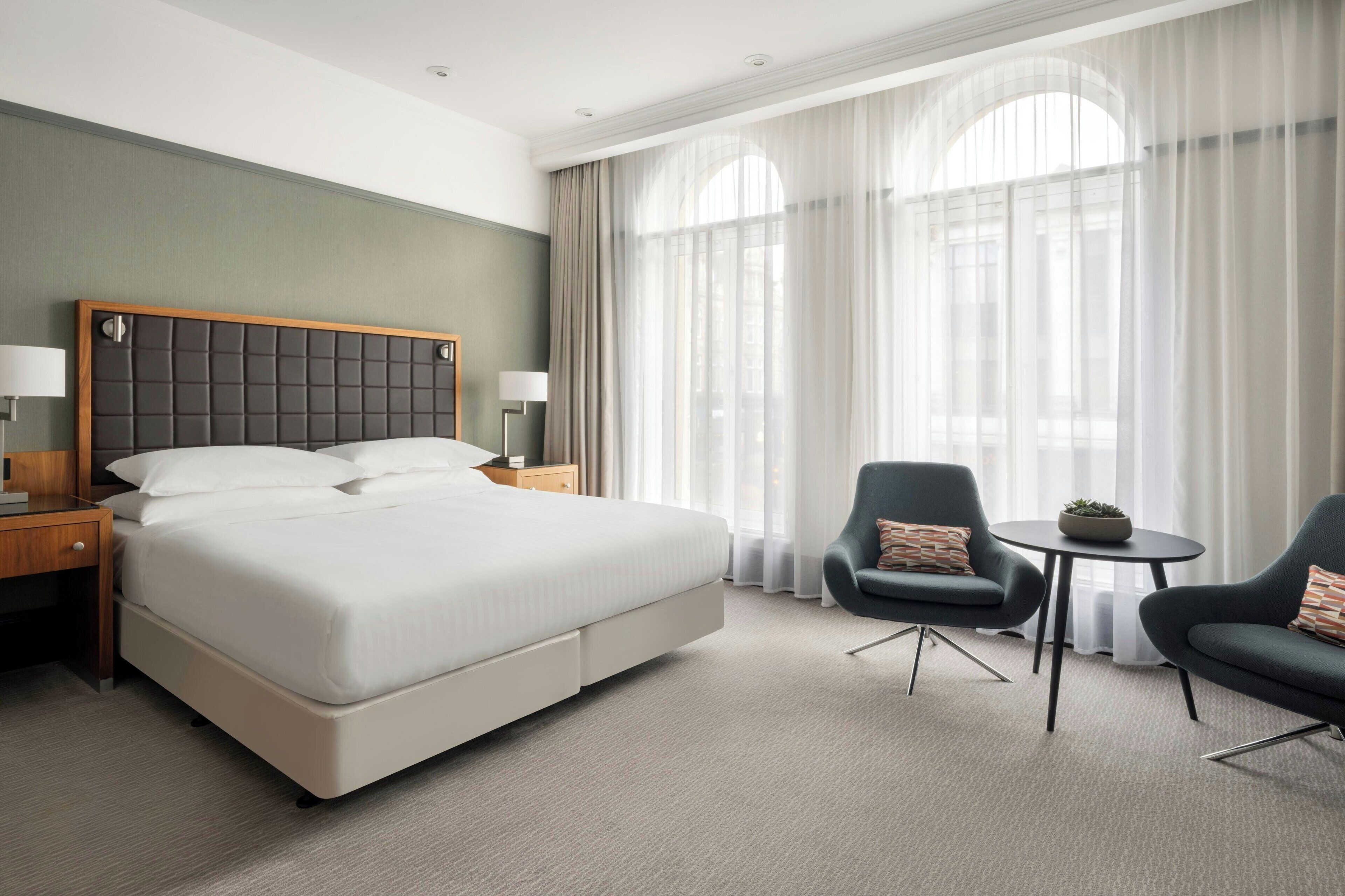 A modern hotel room with a large bed, two armchairs, a small table, and a window with sheer curtains.