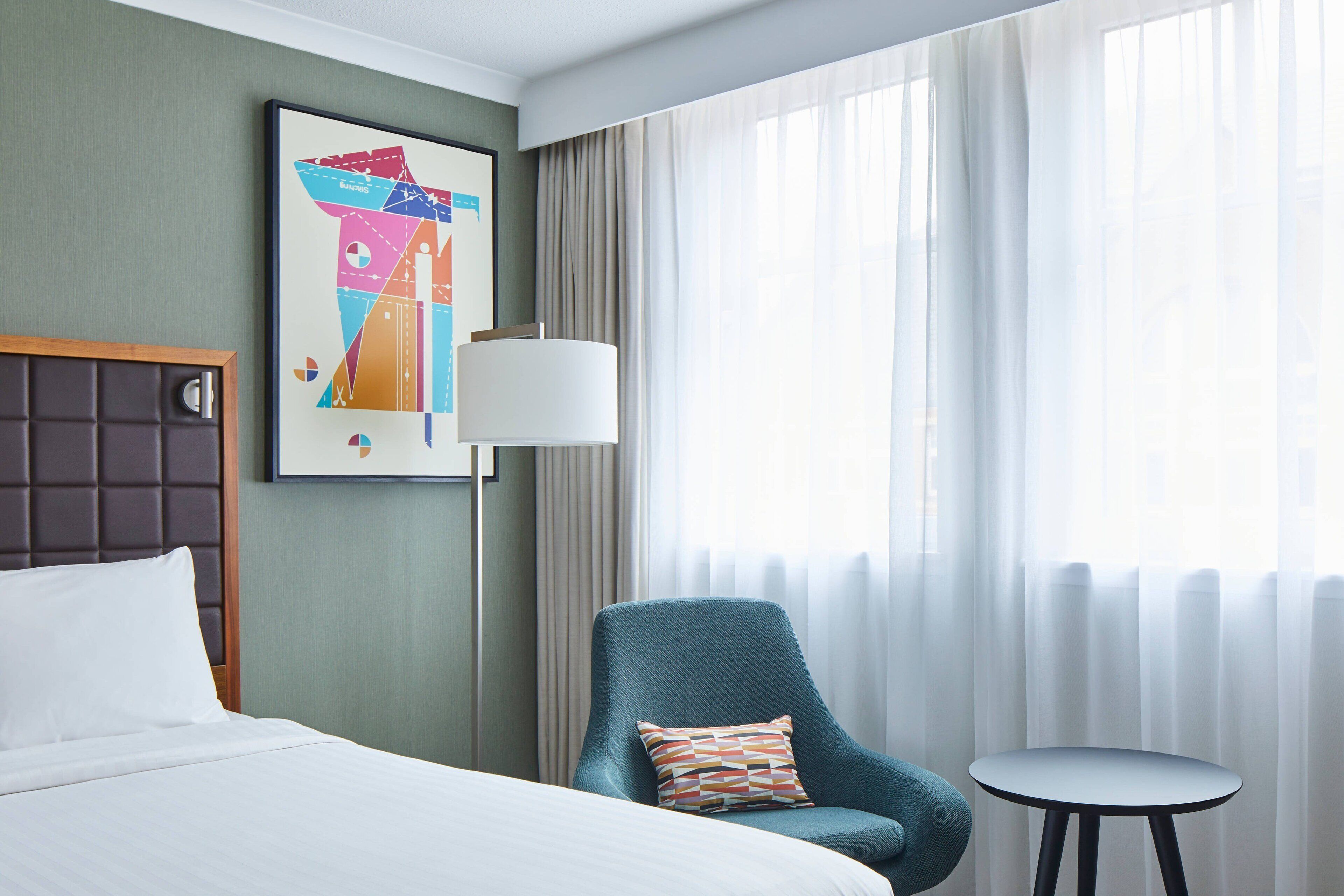 Een hotelkamer met een bed, een stoel, een klein tafeltje, een lamp en een raam met gordijnen.