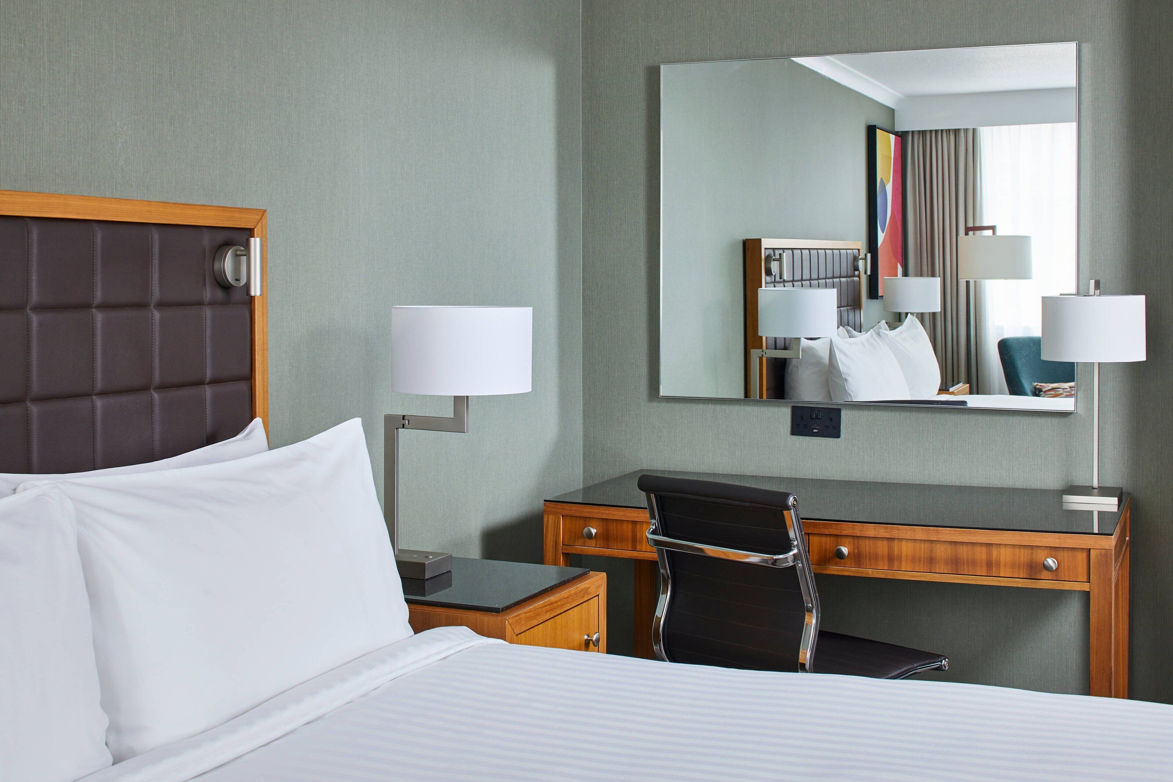 A hotel room with a bed, a desk, a chair, a desk lamp, a bedside table, and a large mirror.