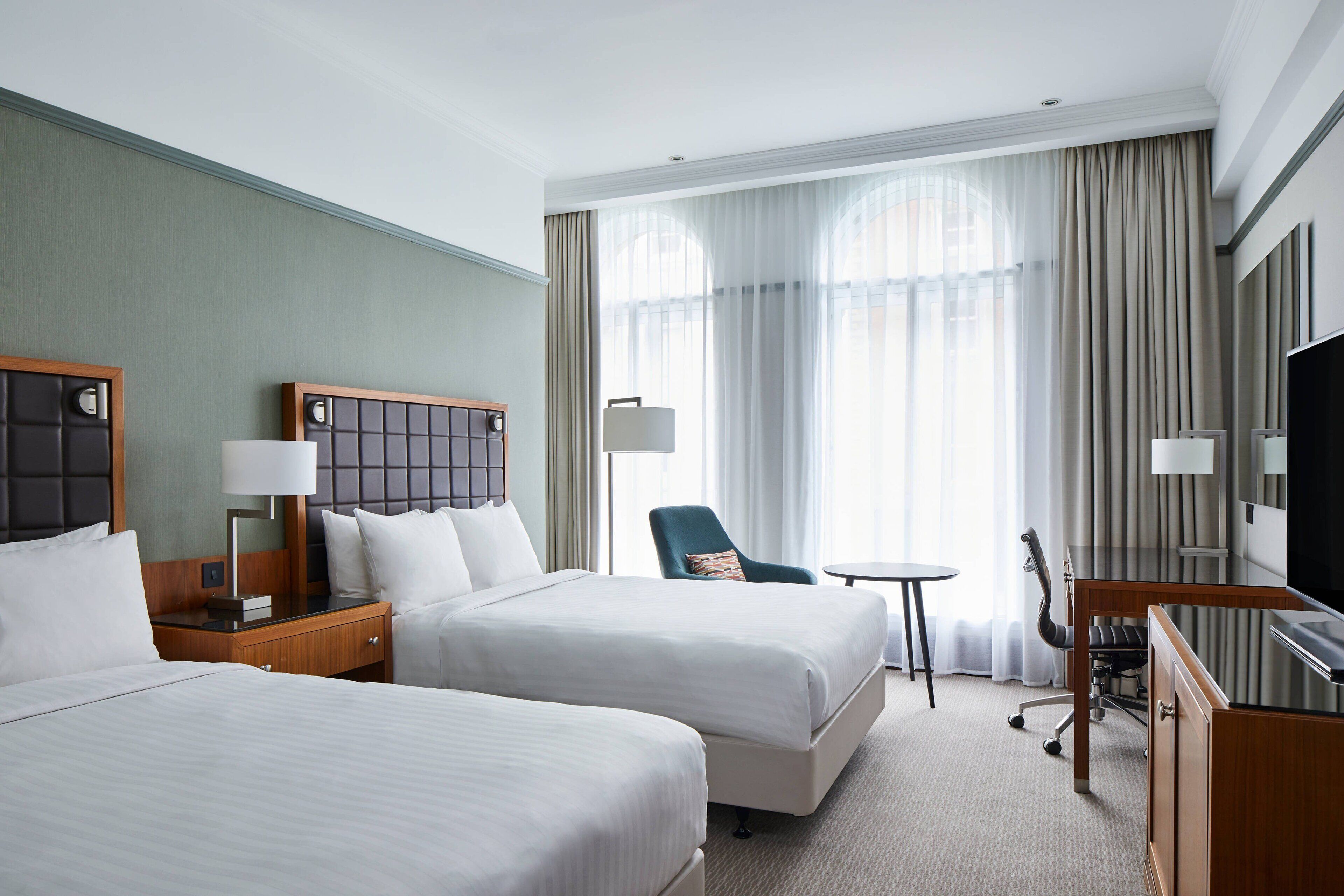 A hotel room with two beds, a desk, a chair, a television, and a large window with curtains.