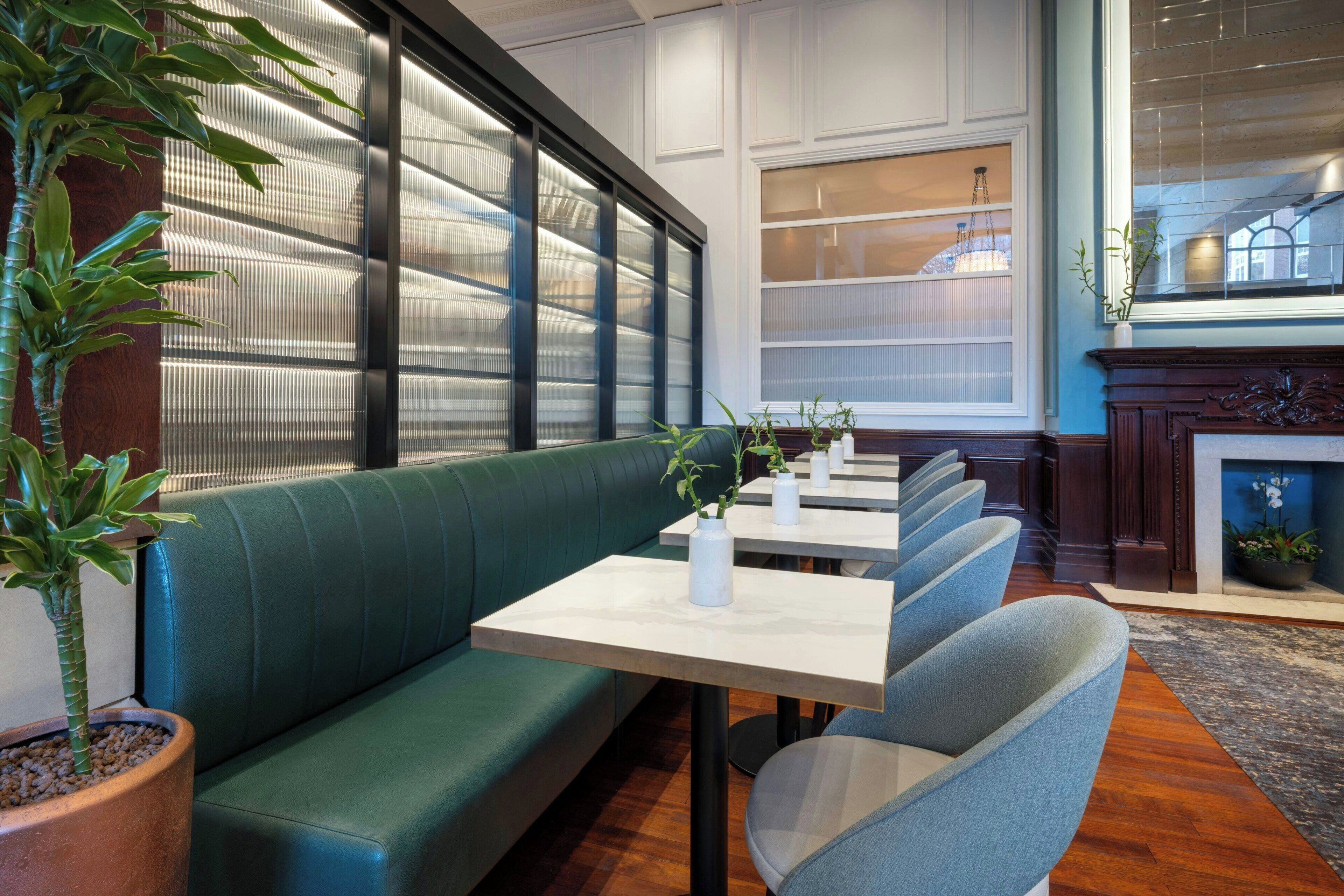 A restaurant interior with green cushioned seating, white tables, and potted plants.