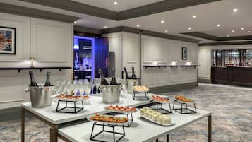 A banquet hall with a long table set for a buffet, featuring various food items and a bar area in the background.