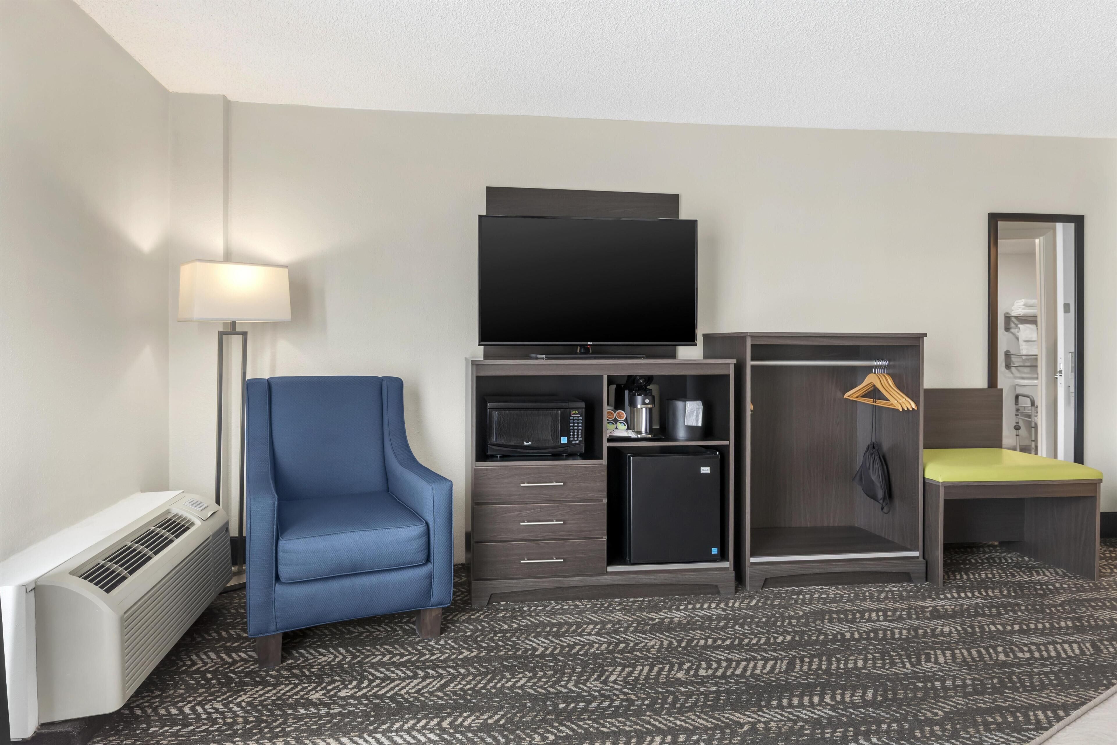 A hotel room with a flat-screen TV, a microwave, a refrigerator, a blue chair, a desk, a hanging coat rack, and a lamp.