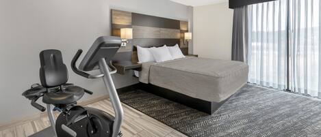 Habitación de hotel con cama, equipo de ejercicio, un ventanal con cortinas y un cabecero moderno.