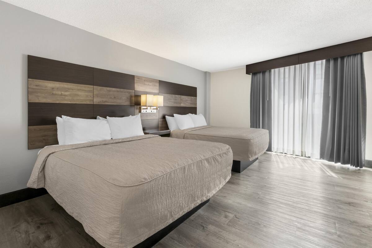 Ein Hotelzimmer mit zwei Betten, einem großen Fenster mit Vorhängen, einem Holz-Kopfteil und einer Wandleuchte.