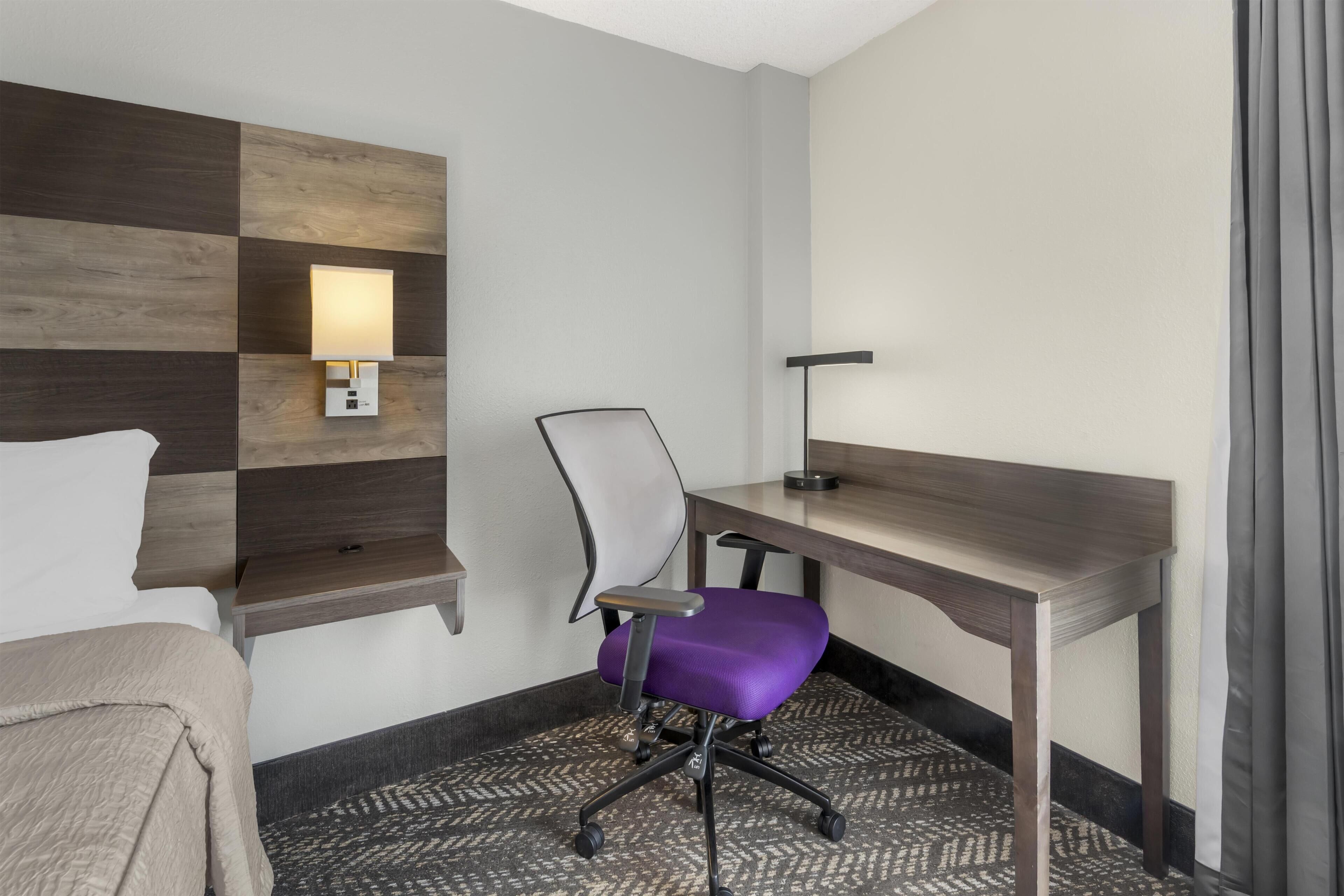 Habitación de hotel con cama, escritorio, silla, lámpara de pared y alfombra estampada.
