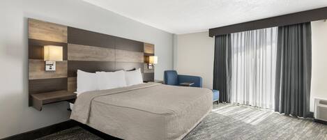 Habitación de hotel con cama, lámparas de noche, escritorio, silla y una ventana grande con cortinas.