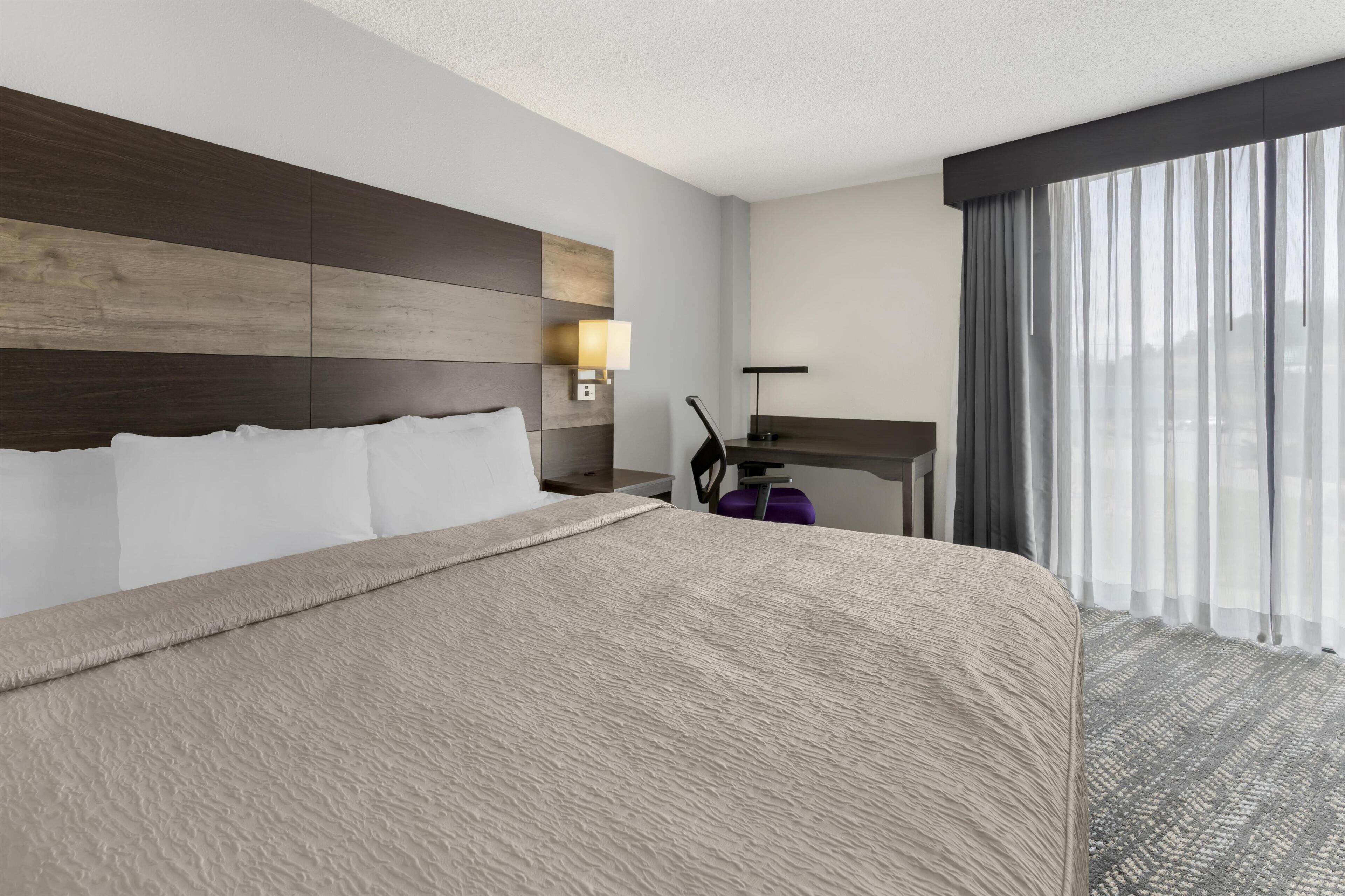 Une chambre d’hôtel avec un grand lit, un bureau, une chaise et une vue sur la ville à travers de fines rideaux.