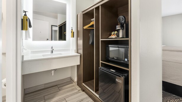 Une salle de bain moderne avec un lavabo blanc, un miroir et un meuble en bois intégrant un four à micro-ondes et un réfrigérateur.