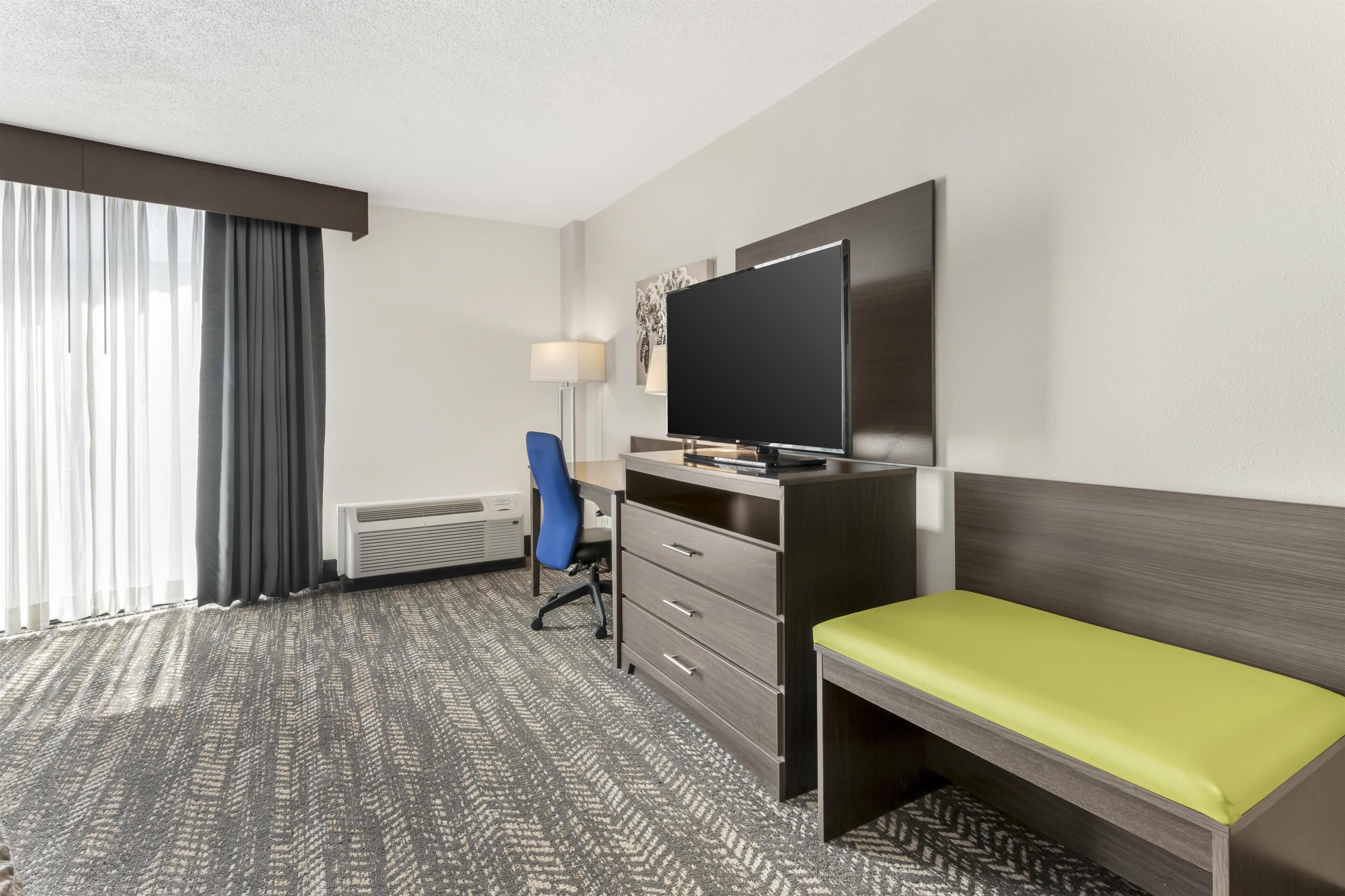 A hotel room with a bed, a desk with a chair, a flat-screen TV, a heater, and a window with curtains.