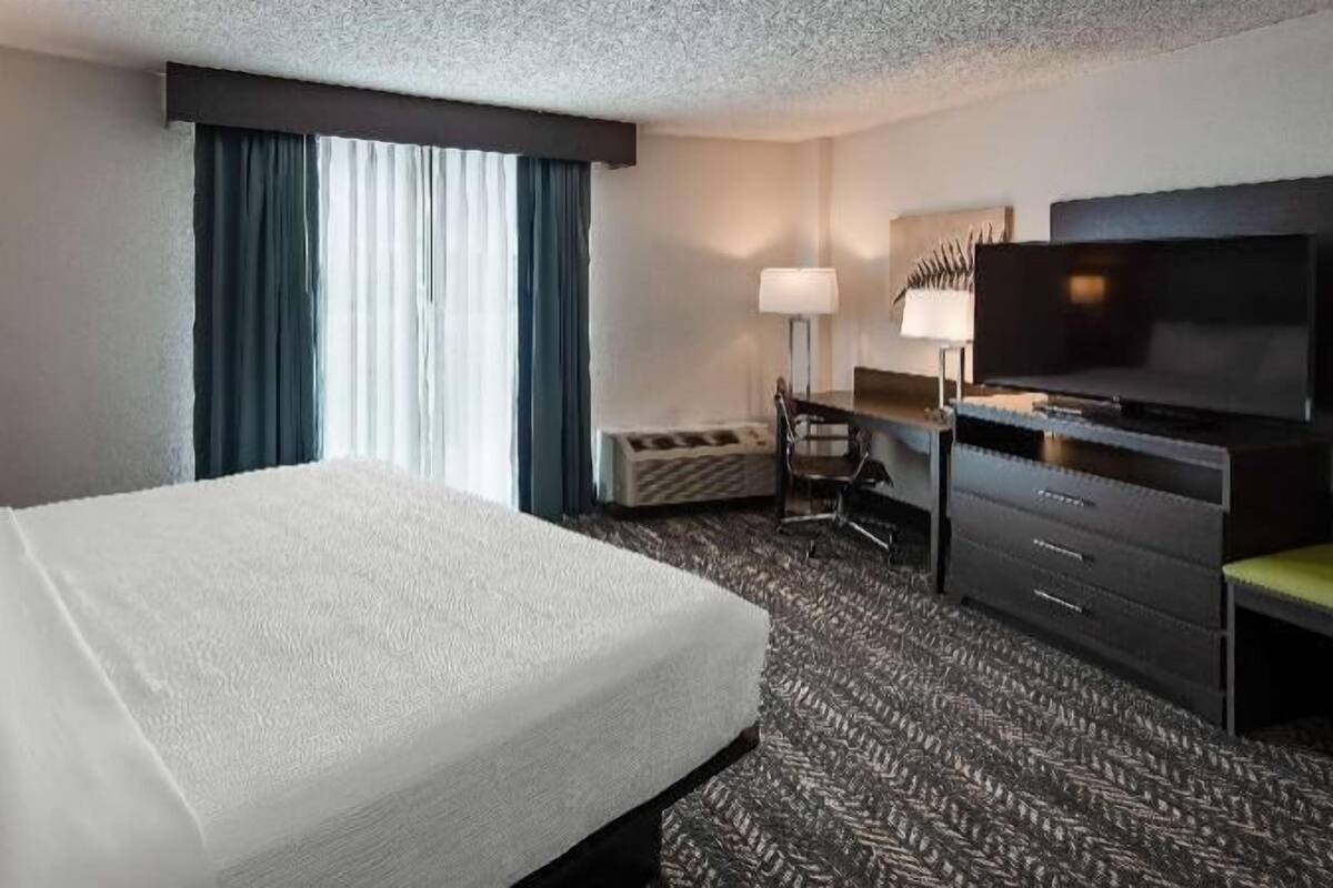 Ein Hotelzimmer mit einem großen Bett, einem Schreibtisch, einem Sessel, einem Fernseher und einem Fenster mit Vorhängen.