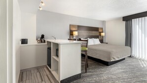Une chambre d’hôtel comprenant un lit, un bureau avec une chaise, une petite kitchenette, une grande fenêtre avec des rideaux et un tapis à motifs.