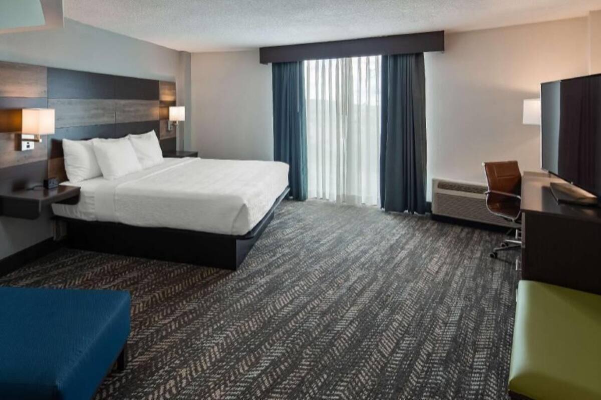 Ein Hotelzimmer mit einem großen Bett, einem Schreibtisch mit Stuhl, einem Fernseher, einem Fenster mit Vorhängen und einem gemusterten Teppich.