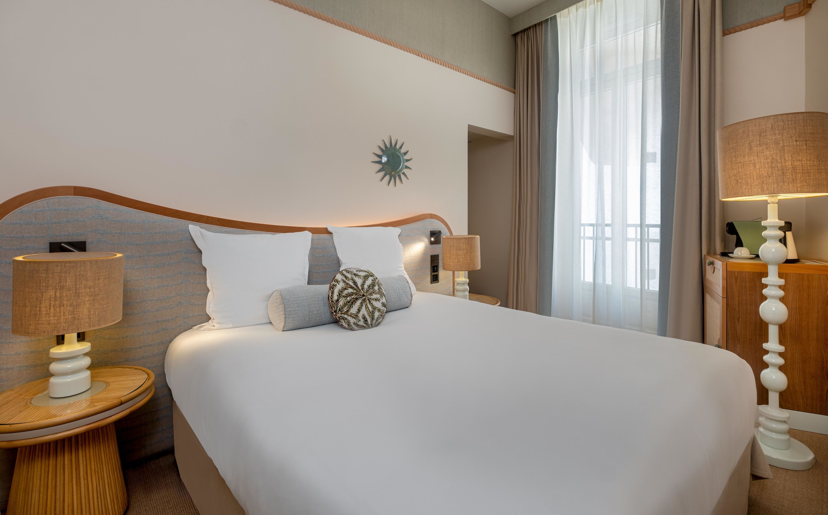 Quarto de hotel com uma cama grande, duas mesas de cabeceira com luminárias, um relógio na parede e uma janela com cortinas.