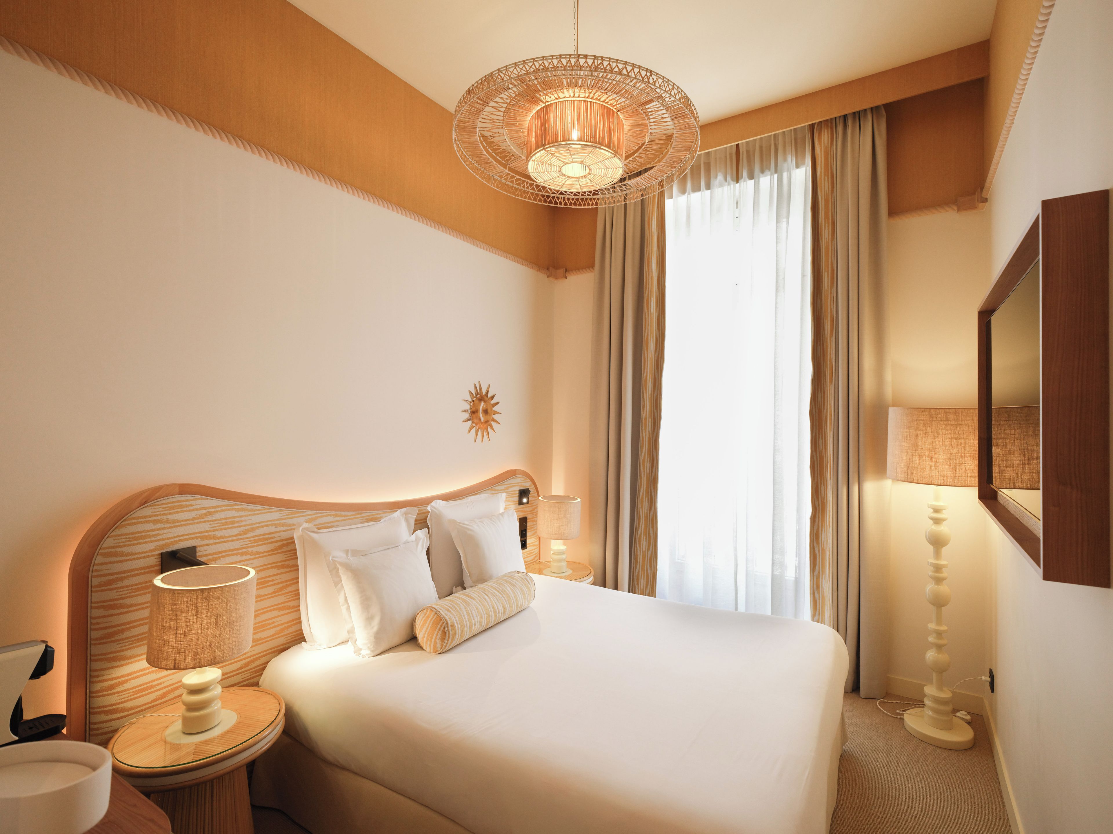 A hotel room with a bed, bedside lamps, a wall-mounted lamp, a mirror, and a window with curtains.
