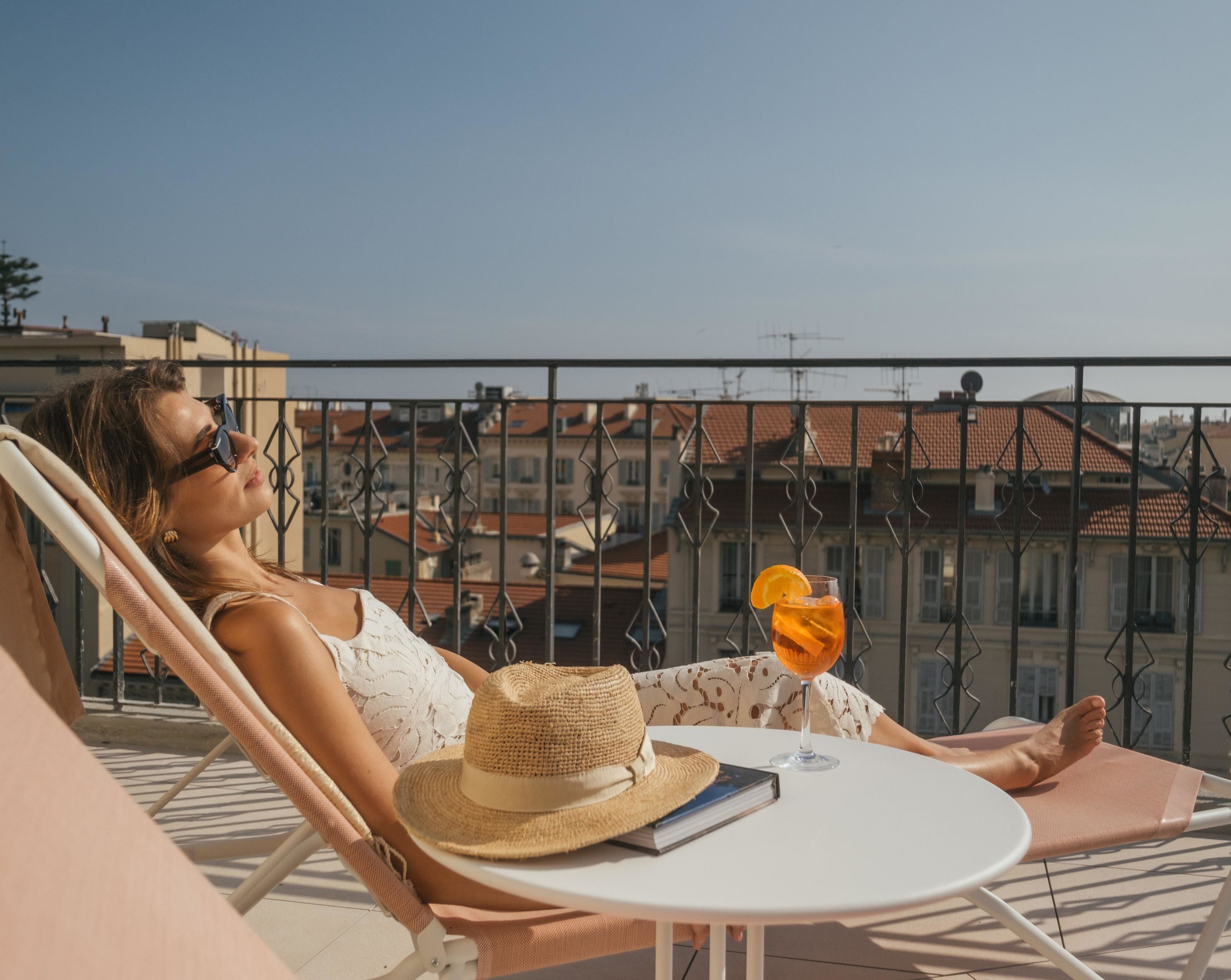 A person reclining on a lounge chair on a balcony with a drink and a hat on the table.