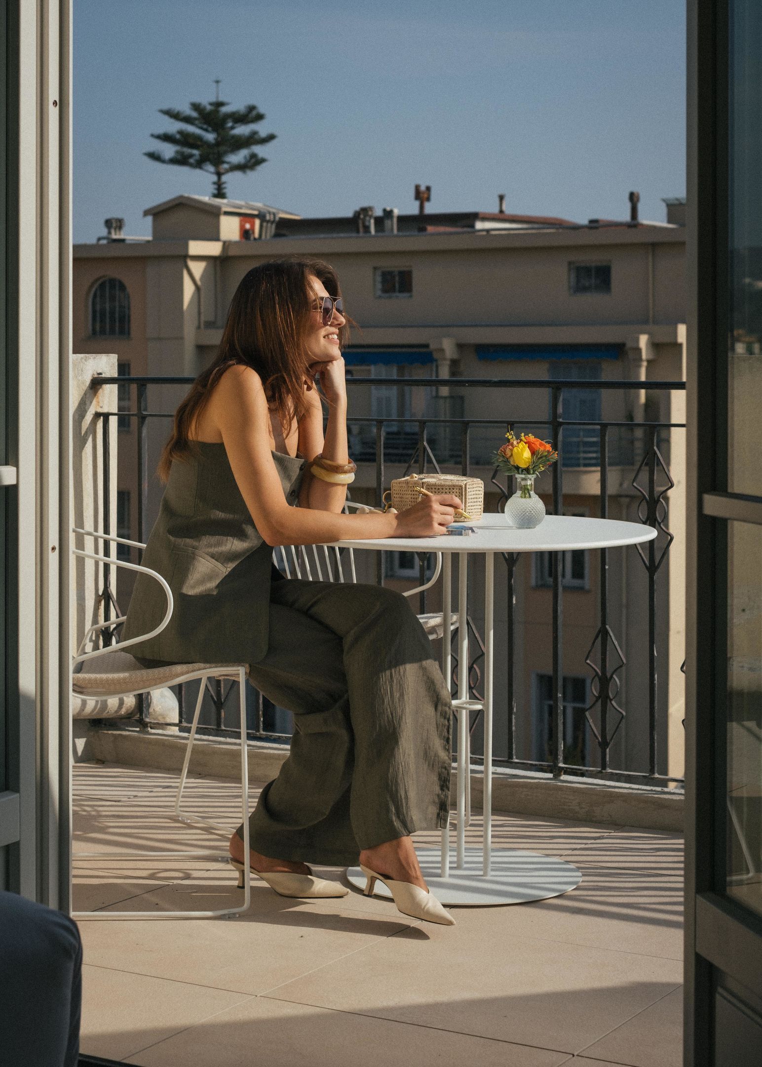 A person seated at a round table on a balcony with a railing, a small vase with flowers, and a building in the background.