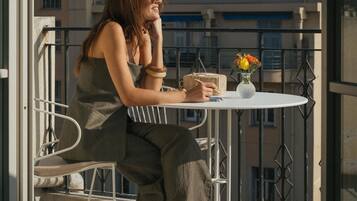 A person seated at a round table on a balcony with a railing, a small vase with flowers, and a building in the background.