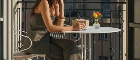A person seated at a round table on a balcony with a railing, a small vase with flowers, and a building in the background.