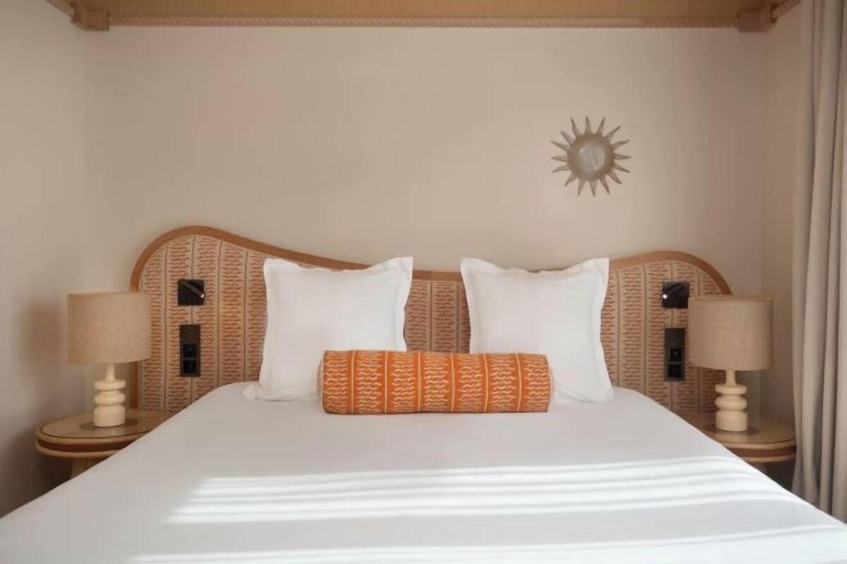 Een net opgemaakt bed met witte lakens en twee kussens, een wanddecoratie in de vorm van een zon, en twee nachtlampjes op houten nachtkastjes.