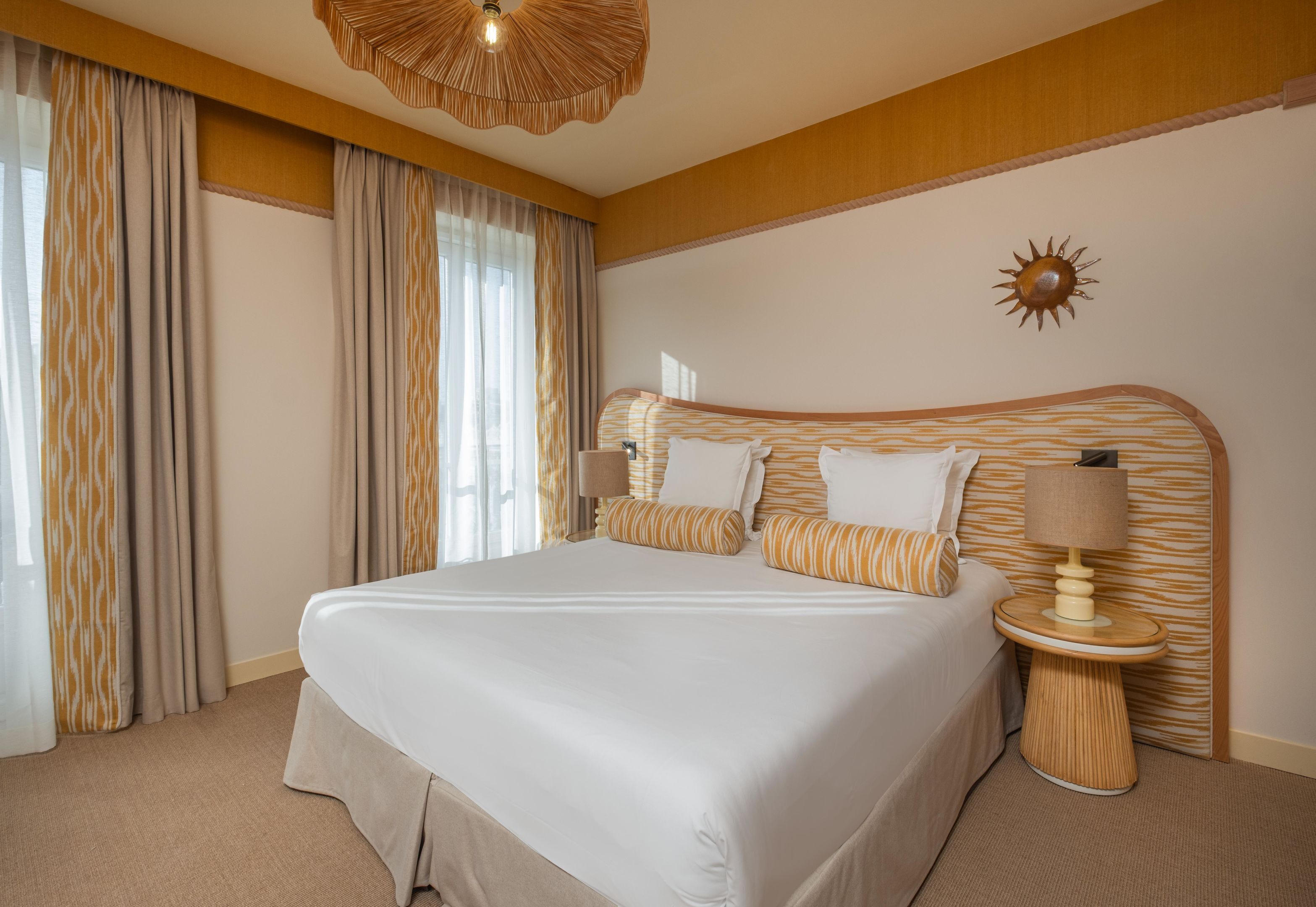 A hotel room with a large bed, a bedside table with a lamp, and a sun decoration on the wall.