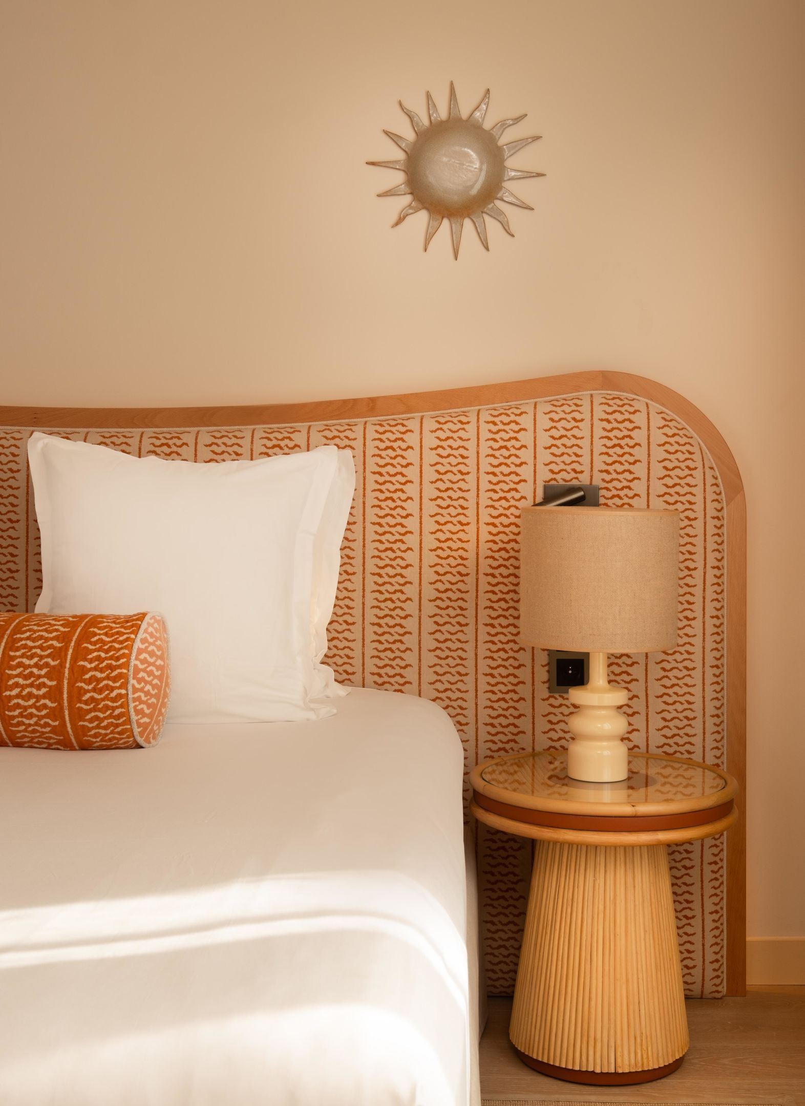 A neatly made bed with white linens and two pillows, a wall-mounted lamp beside it, and a patterned wallpaper.