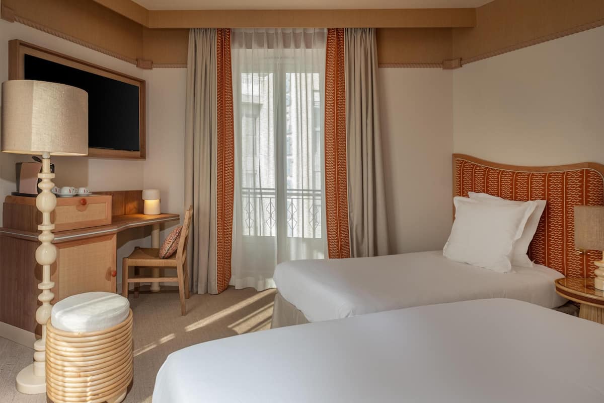 A hotel room with two beds, a desk with a lamp, a chair, and a large window with curtains.