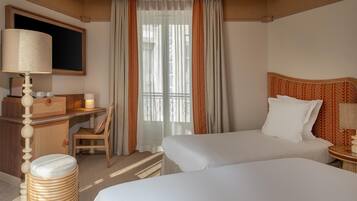 Habitación de hotel con dos camas, un escritorio con lámpara, una silla y un amplio ventanal con cortinas.