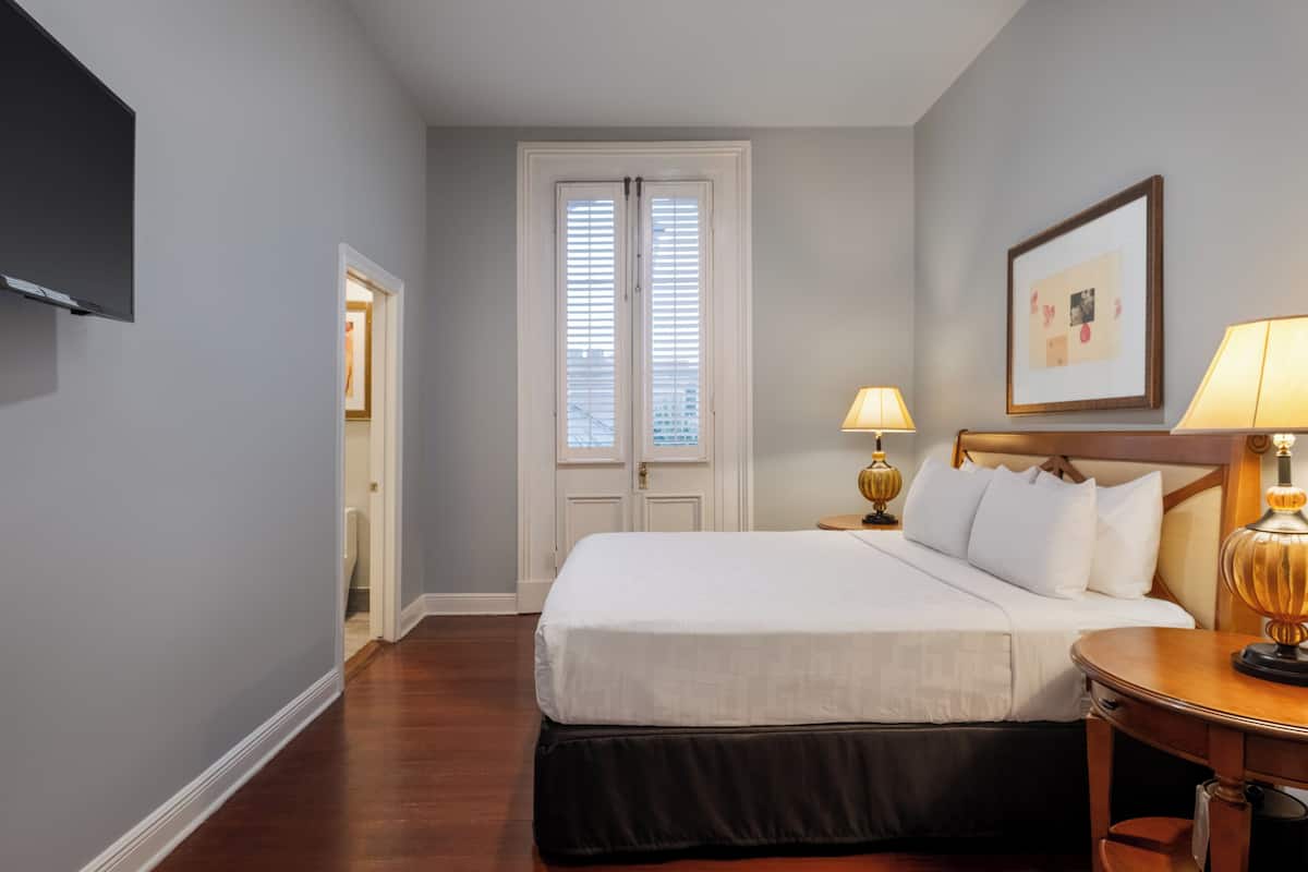 A hotel room with a bed, bedside table with lamps, a framed picture on the wall, and a door with blinds.