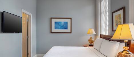 A hotel room with a bed, a flat-screen TV mounted on the wall, a framed abstract painting, two bedside lamps, and a window with blinds.