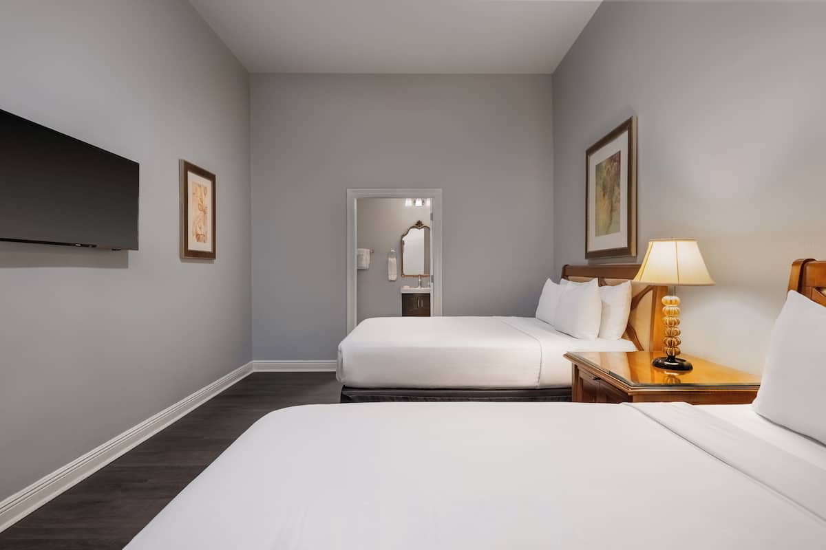 A hotel room with two beds, a TV mounted on the wall, a bedside table with a lamp, and framed artwork on the walls.