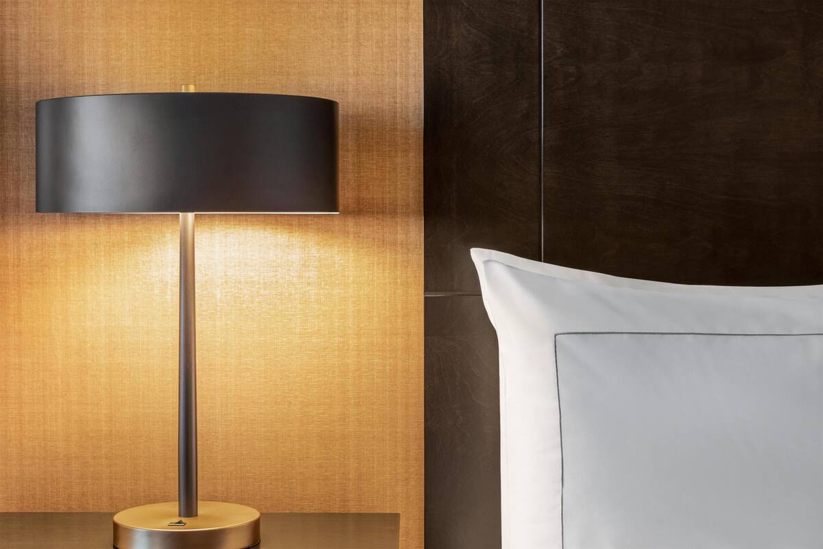 Eine moderne Lampe mit zylindrischem Lampenschirm, eine Wand mit Holzstruktur und ein weißes Kopfkissen auf einem Bett.