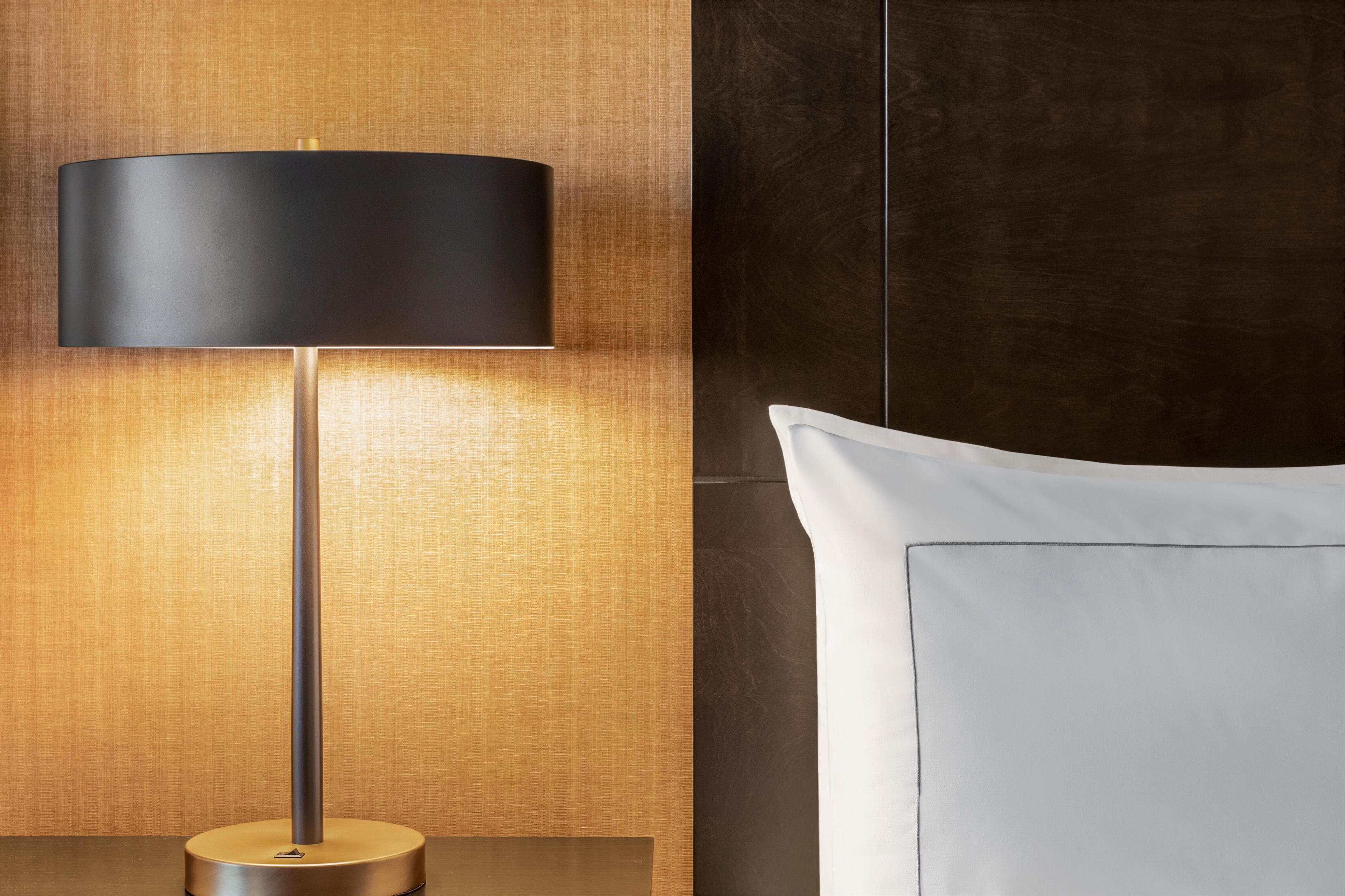 A modern lamp with a cylindrical shade, a wooden textured wall, and a white pillow on a bed.