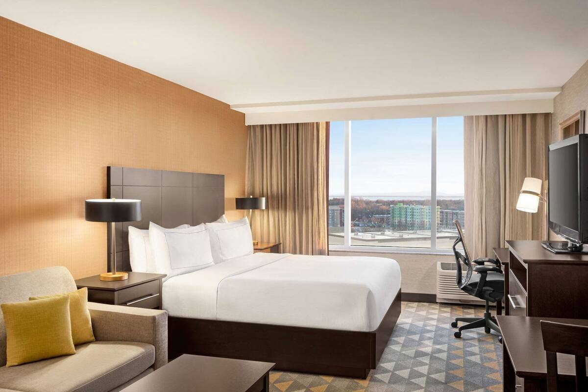 Ein Hotelzimmer mit einem großen Bett, einem Schreibtisch, einem Sessel, einem Fernseher und Blick auf Gebäude durch das Fenster.