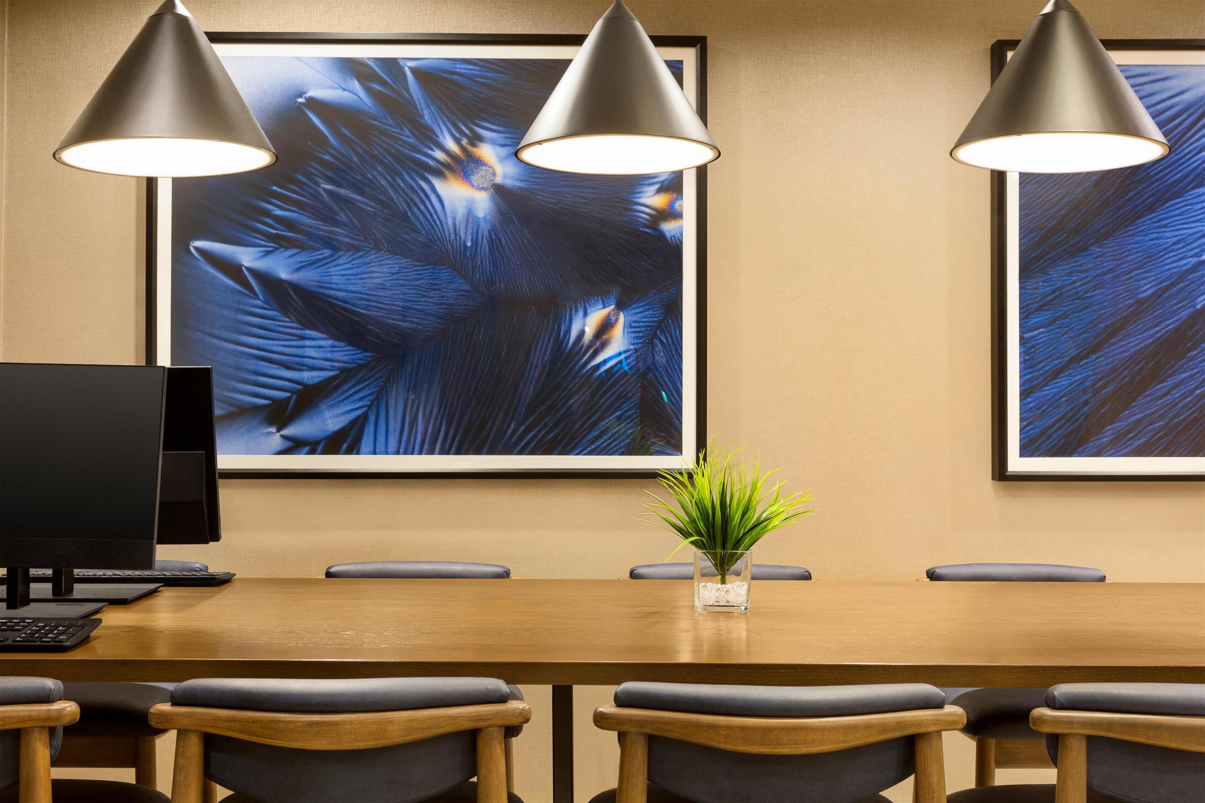 A conference room with a long table, six chairs, two pendant lights, a computer, and a framed abstract painting on the wall.