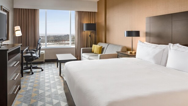 Een hotelkamer met een groot bed, een bureau met stoel, een bank, een televisie en een groot raam met gordijnen.