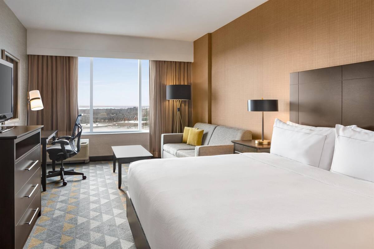 Ein Hotelzimmer mit einem großen Bett, einem Schreibtisch mit Stuhl, einer Couch, einem Fernseher und einem großen Fenster mit Vorhängen.